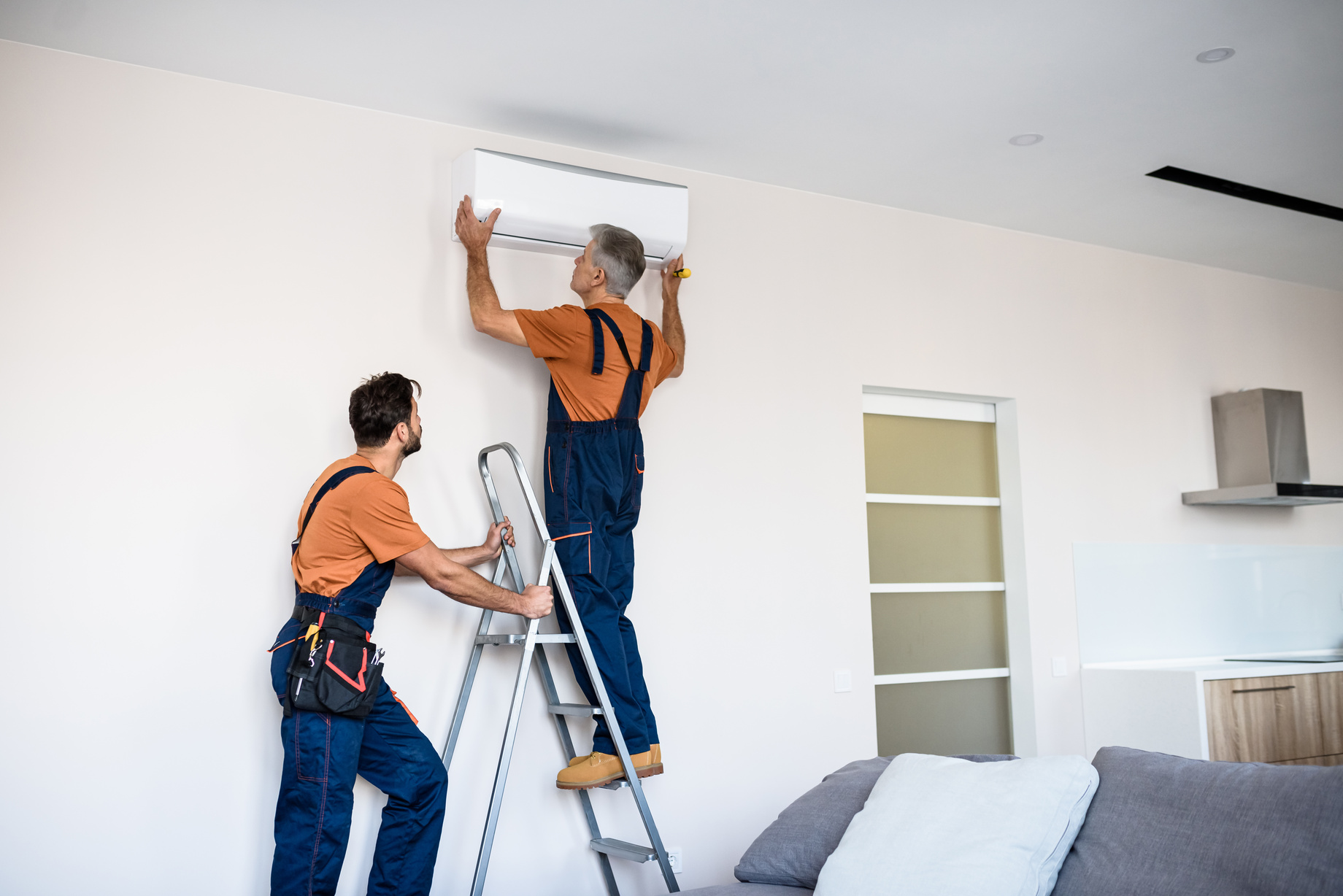 Two workers in uniform, using ladder while installing a new air conditioner in the apartment.