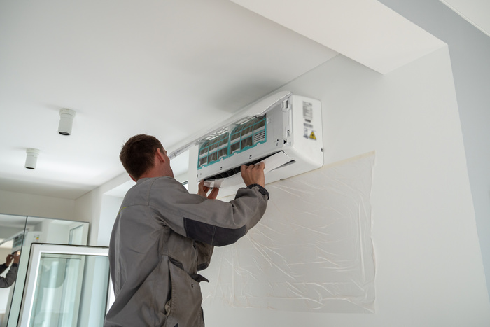Male worker wearing uniform repairing air conditioner in apartment during summer season, man technician standing indoors repairing HVAC system, checking and replacing AC filter